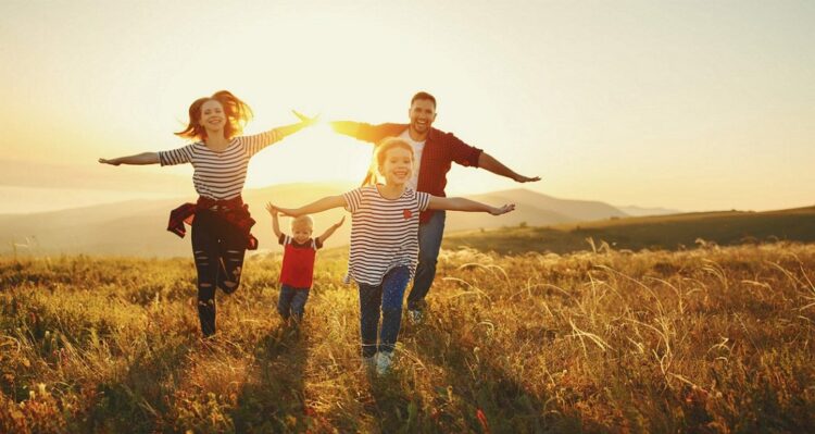 Frühbucherrabatt Urlaub 2024 Familie bei REISEBÜRO Wache; im Bild: Frau, Mann, 2 Kinder, die durch ein Feld rennen, die Arme ausgebreitet, hinter ihnen geht die Sonne unter