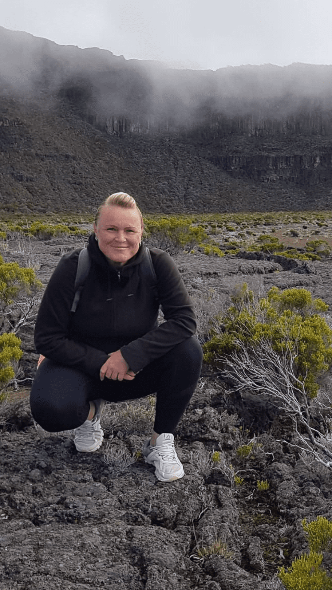 La Réunion Urlaub buchen Aktivurlaub buchen beim REISEBÜRO Wache Erfurt; im Bild: eine blonde Frau, in Outdoor-Kleidung hockt auf einem erkalteten Lavafeld zwischen grünen Pflanzen, im Hintergrund Berge in Rauchwolken