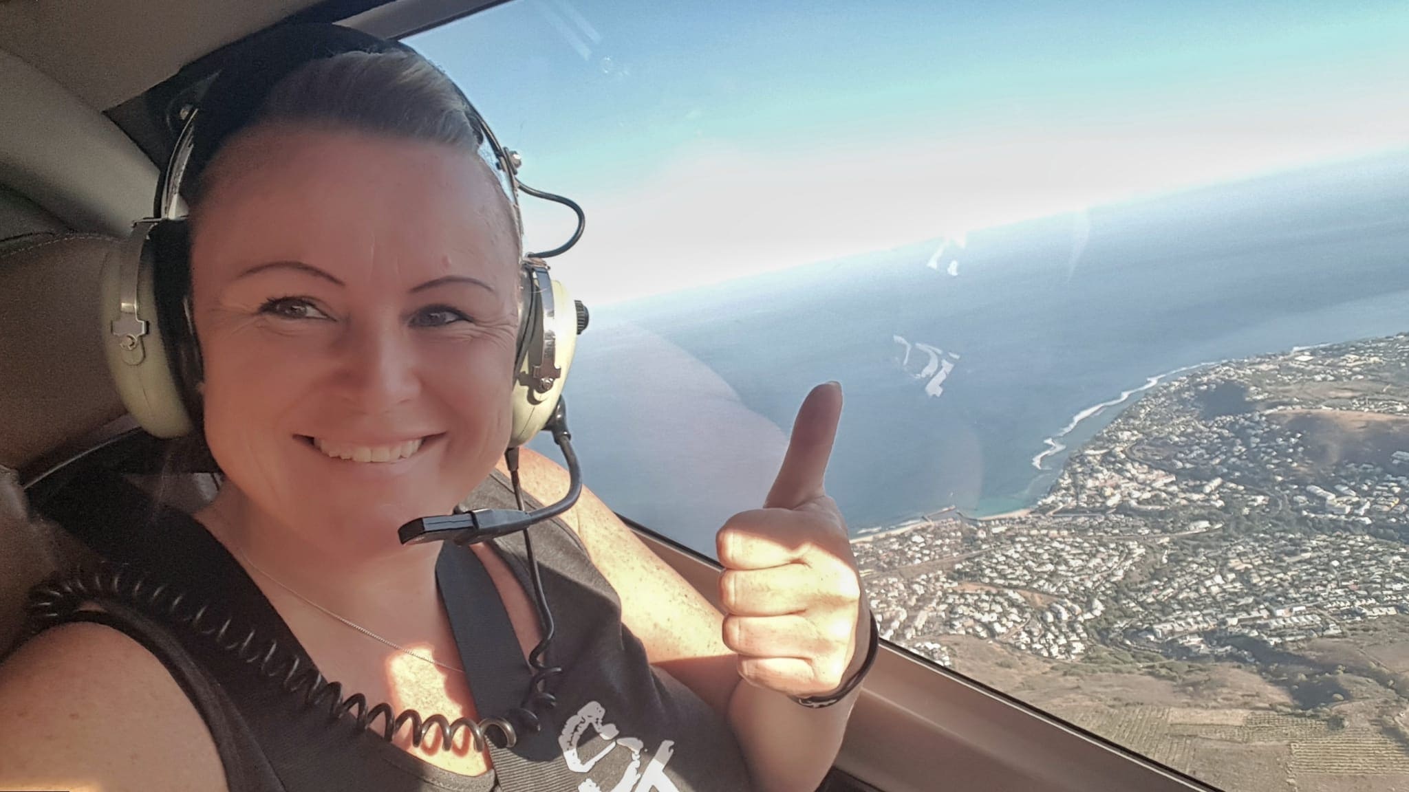 Helikopterflug über La Réunion beim REISEBÜRO Wache in Erfurt buchen; im Bild: Blonde Frau Mitte 40 in einem Flugzeug mit Headset auf, lächelt, streckt den Daumen nach oben; im Hintergrund die Insel und Meer