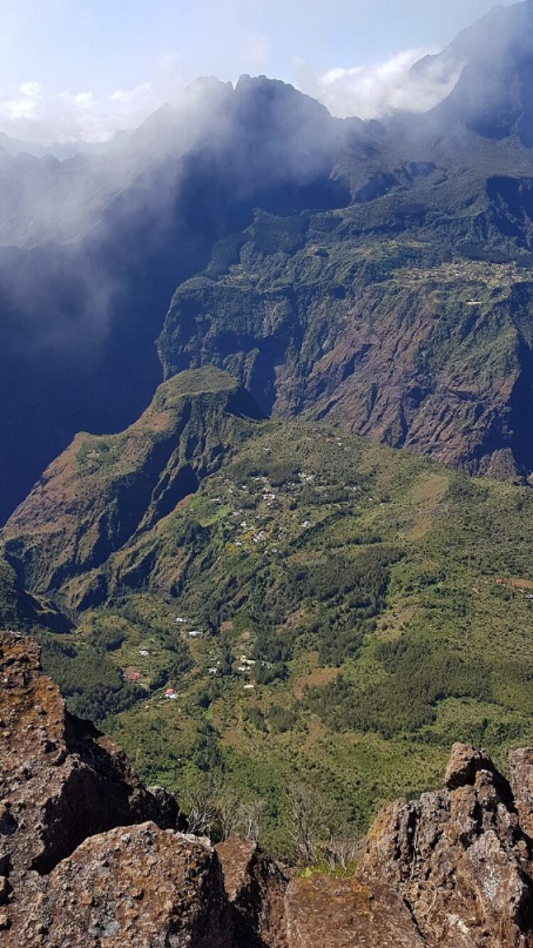 La Reunion Urlaub buchen beim REISEBÜRO Wache in Erfurt; im Bild: Blick vom Gipfel auf La Reunion, im Vordergrund bräunliches Gestein, grün bewachsen, in der Ferne Gipfel von Wolken oder Rauch verhangen