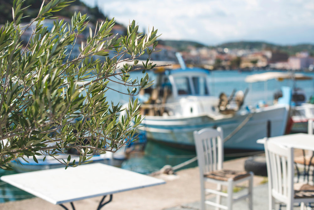 Griechenland Urlaub buchen im Erfurter REISEBÜRO Wache; im Bild: weiße Restaurant-Tische am Hafen in Griechenland, im Hintergrund ein Boot