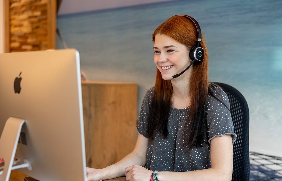 Job Reisebüro Erfurt? Stellen Reisebüro Erfurt? - Hier finden Sie ihn: beim REISEBÜRO Wache in Erfurt; im Bild: junge Frau mit langen, rötlichen Haaren telefoniert über ein Headset, während sie an einem Bildschirm arbeitet