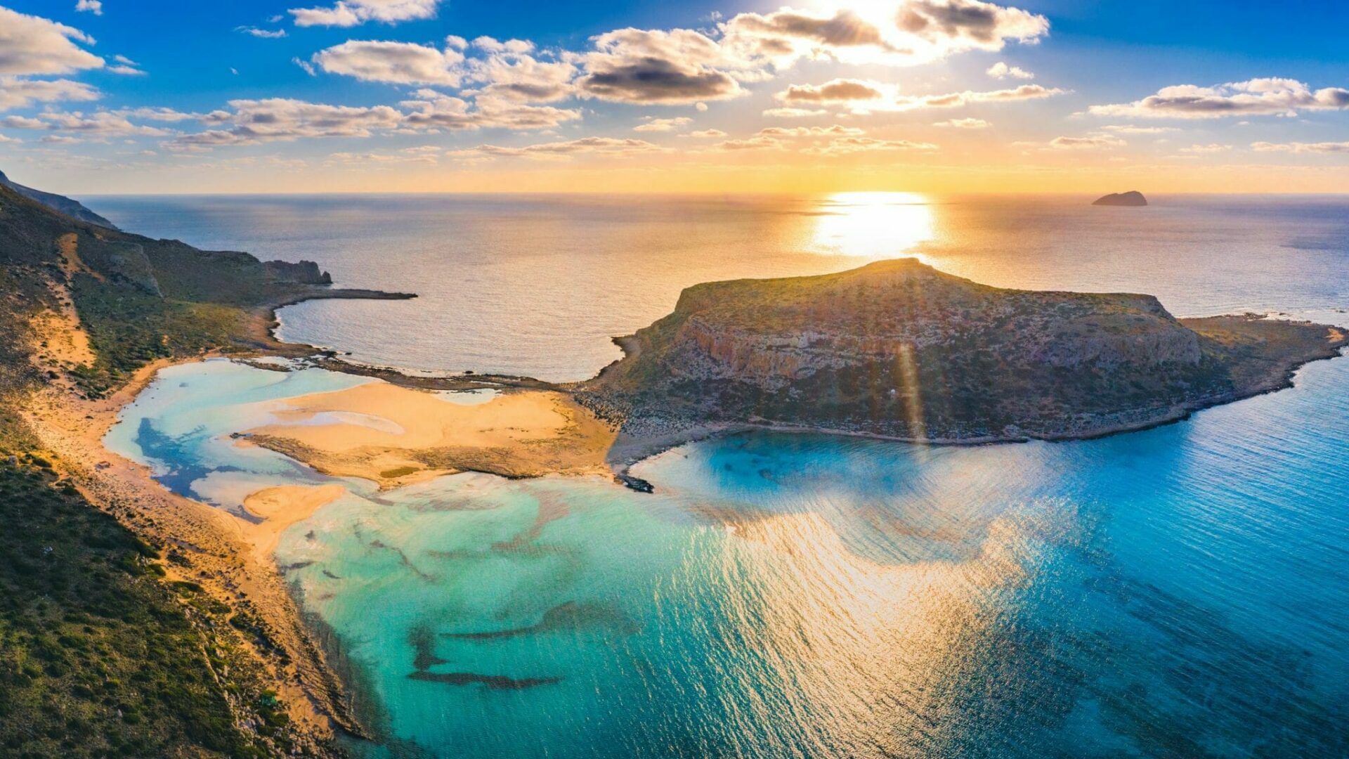 Urlaub Griechenland 2025 buchen beim REISEBÜRO Wache, Erfurt; im Bild: Felsen, die mit einer Sandbank-Verbindung mit einem Felsen im Meer verbunden sind, die Sonne versinkt dahinter im Meer