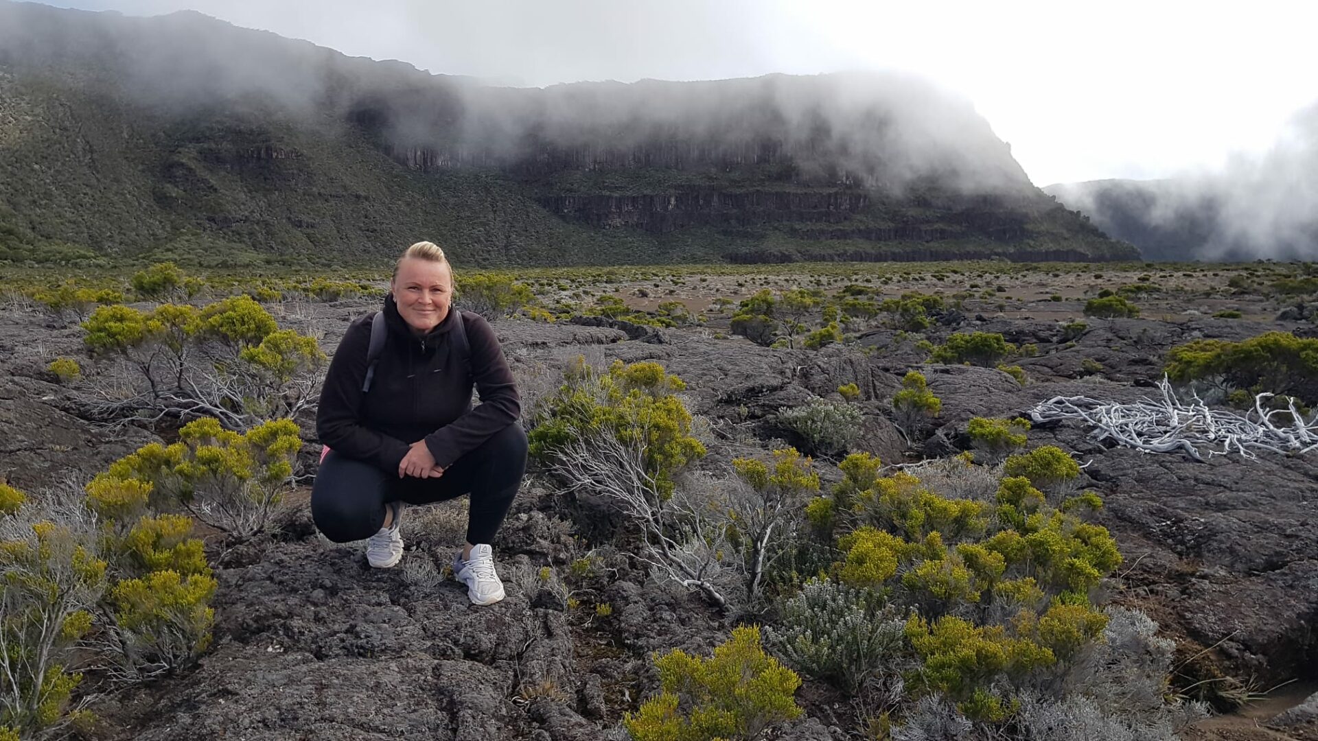 La Réunion Urlaub buchen Aktivurlaub buchen beim REISEBÜRO Wache in Erfurt; im Bild: Reiseverkehrskauffrau Anke Fröhlich, blond, in schwarzer Kleidung, weißen Turnschuhen, hockt auf einem erkalteten Lavafeld zwischen flachen grünen Pflanzen, im Hintergrund Berge in Rauchwolken